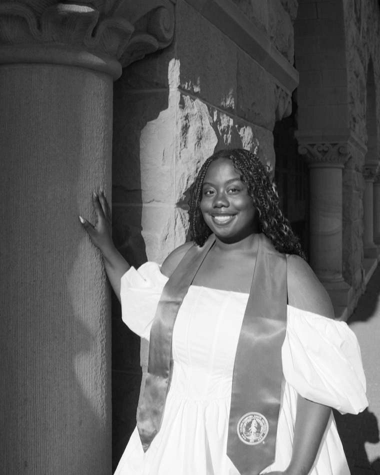 Nyah Ware in a white dress and Stanford graduation stole, standing beside a sandstone column